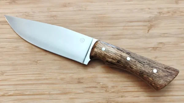 Full left view of the Bur Oak Camper/Chopper knife featuring a 6 1/4" brushed clip point blade crafted from 1095 high carbon steel and a stabilized Florida bur oak handle with mild steel pins, resting on a wooden surface.