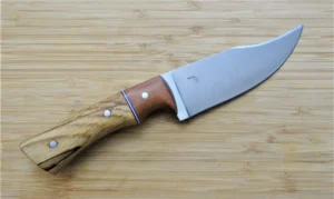 Pecan Nada Bowie large camp knife with upswept clip point blade, Wild Plum bolster, and red, white, blue G10 spacers, right side view on wooden surface.