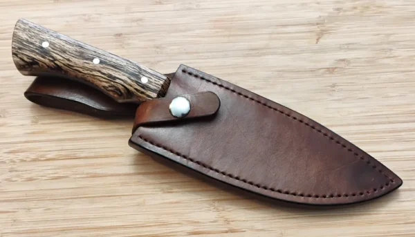 Bur Oak Camper/Chopper knife sheathed in its custom handcrafted leather sheath with a retention strap, showing the stabilized Florida bur oak handle and part of the 1095 high carbon steel blade, on a wooden background.