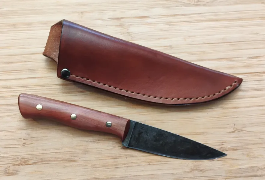 Red Cedar Bird and Trout Knife next to custom hand cut and stitched leather sheath, on wood.