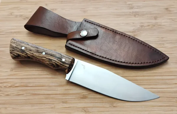 Bur Oak Camper/Chopper knife placed next to its custom handcrafted leather sheath with a retention strap, displaying the 6 1/4" brushed clip point blade and stabilized Florida bur oak handle on a wooden surface.