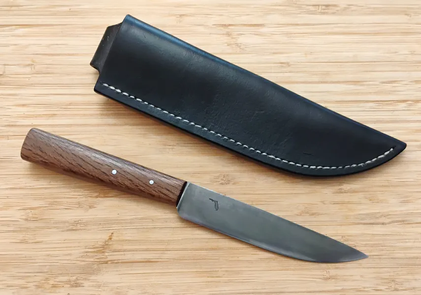 Live Oak French Trade Knife #361 next to its black leather sheath with white linen stitches on a wooden surface