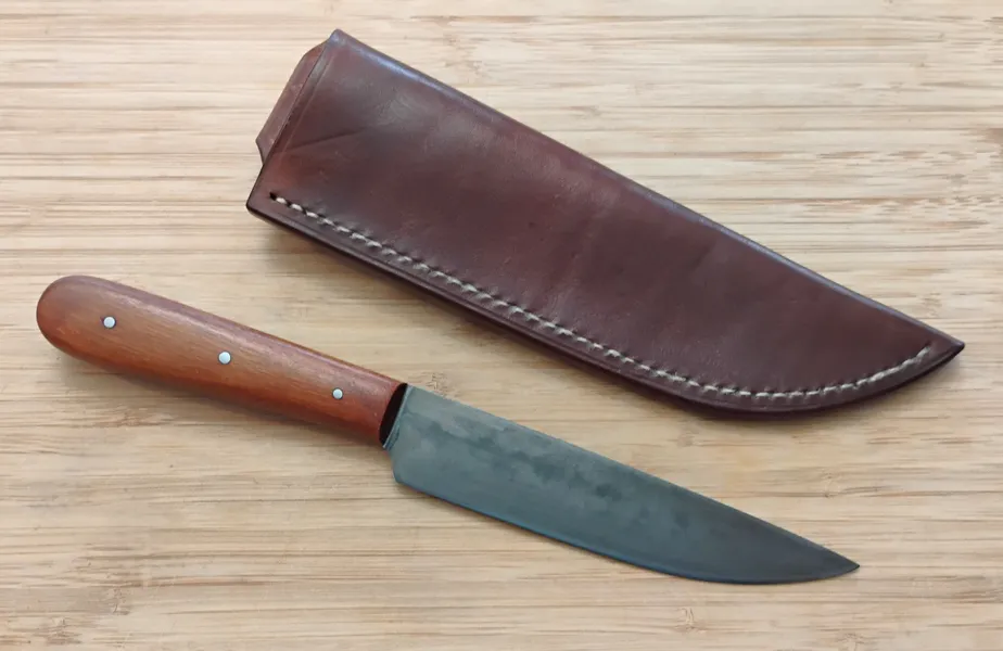 Pernambuco French Trade Knife next to its brown Oak Bark leather sheath with white linen stitches on a wooden surface