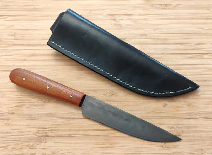 Pernambuco Trade Knife sitting next to a Vinegaroon Black sheath with white stitching on a wooden background.