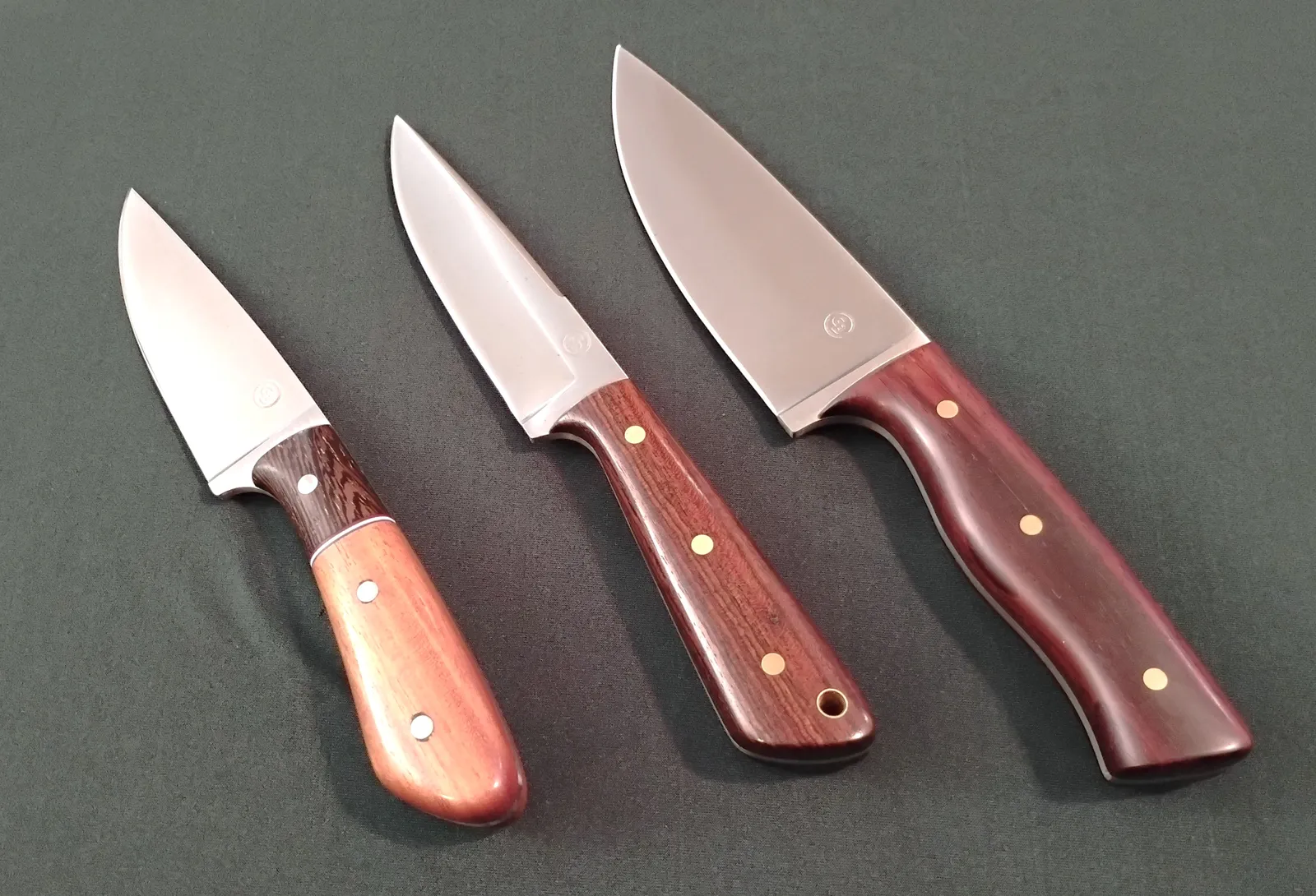 Three custom knives with various exotic wood handles arranged from small to largest with different features on a green background