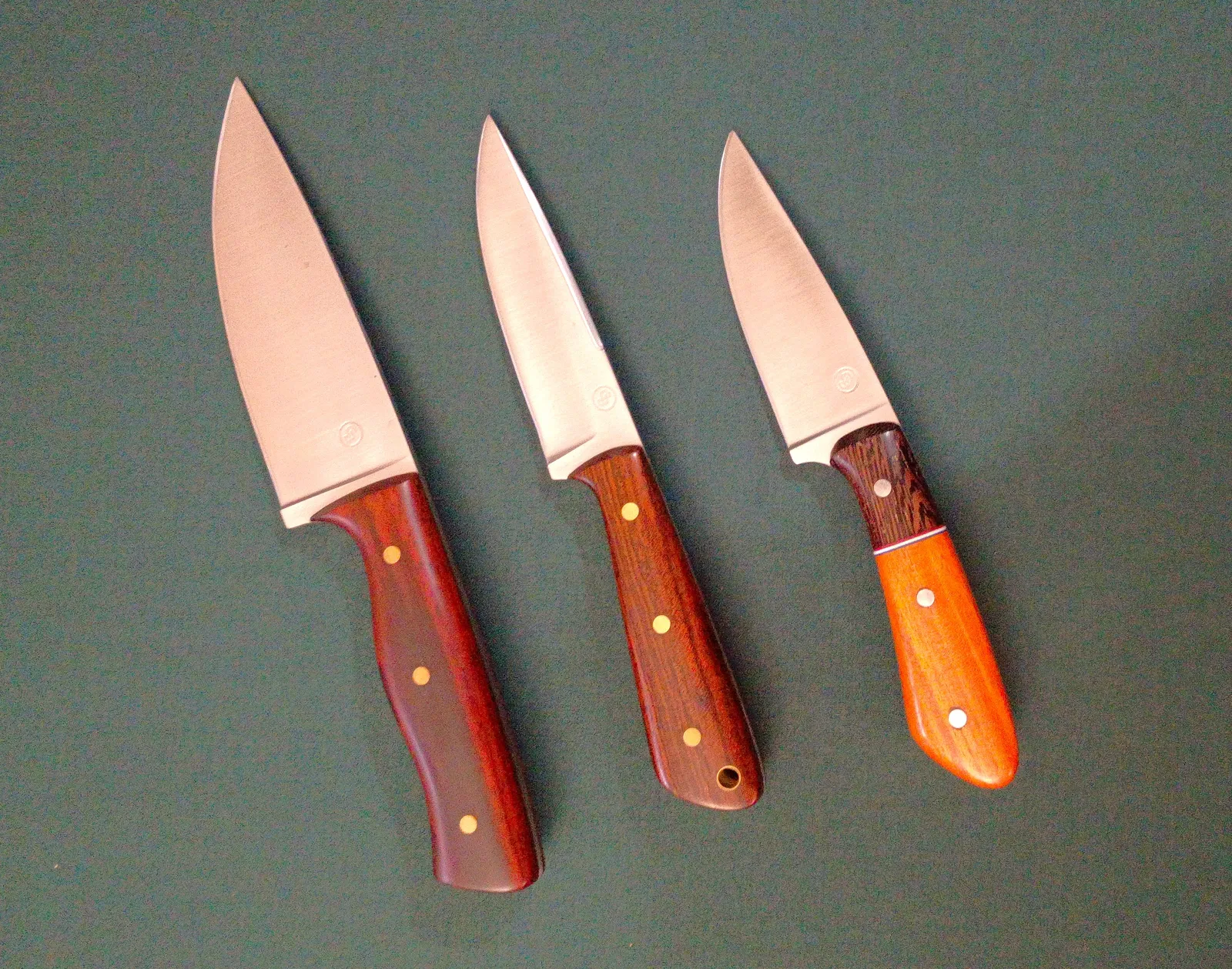 Three custom knives with various exotic wood handles arranged from small to largest with different features on a green background