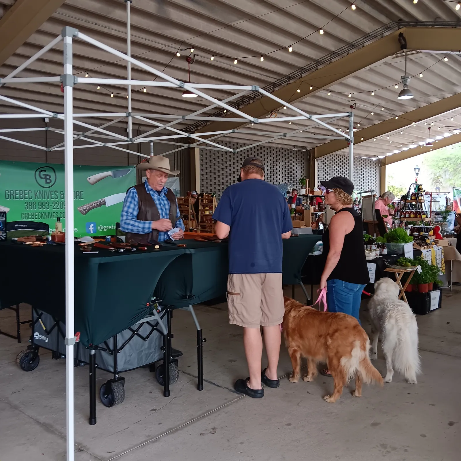 View of GreBec Knives booth at Ocala Downtown Market: Greg talking with two customers with friendly golden retrievers.