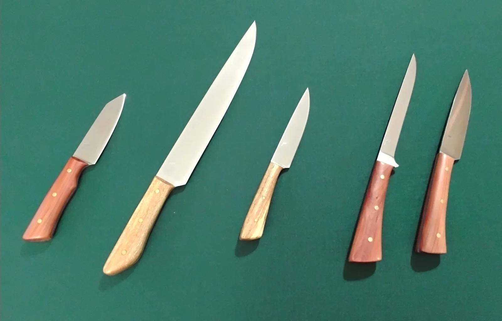 Five custom kitchen knives lined up—paring, utility, boning, carving—with 1095 high carbon steel blades and stabilized Florida wood scales on green backdrop.