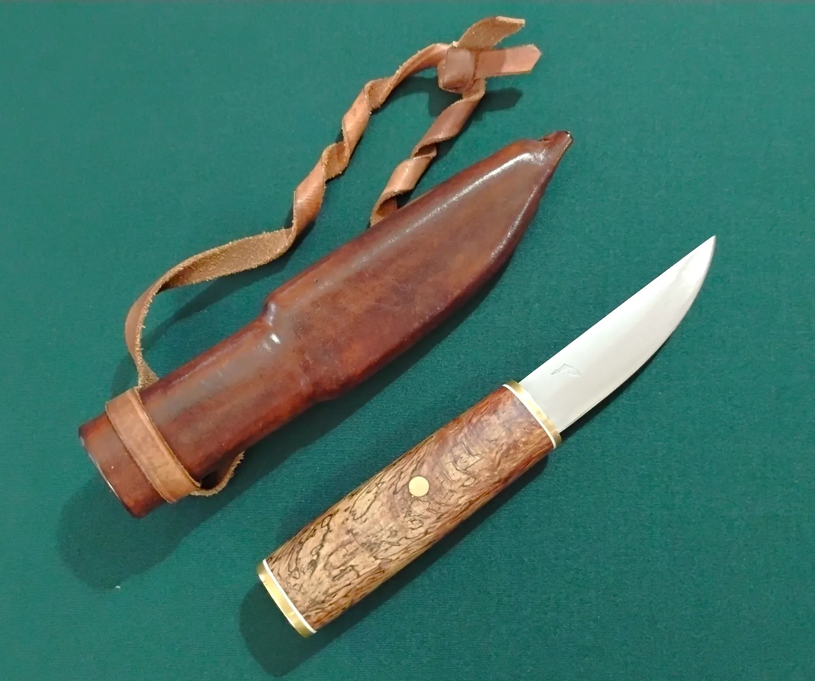 Handcrafted Puukko: stabilized Southern Red Oak handle, brass bolster & pommel, brushed blade, with custom leather sheath on green backdrop.