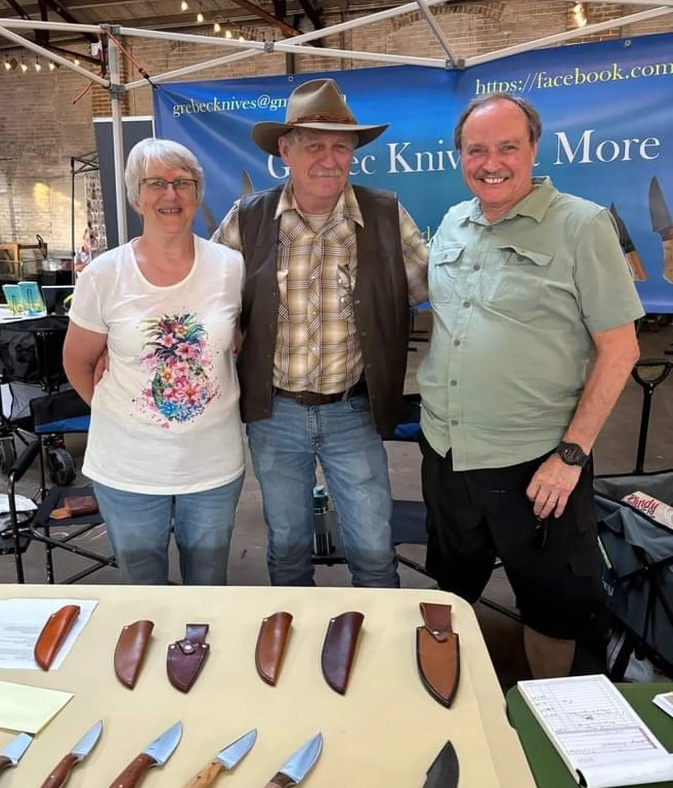 Greg and Becky from GreBec Knives standing with a happy new customer at DeLand Artisans Market with display table with available knives in forefront.
