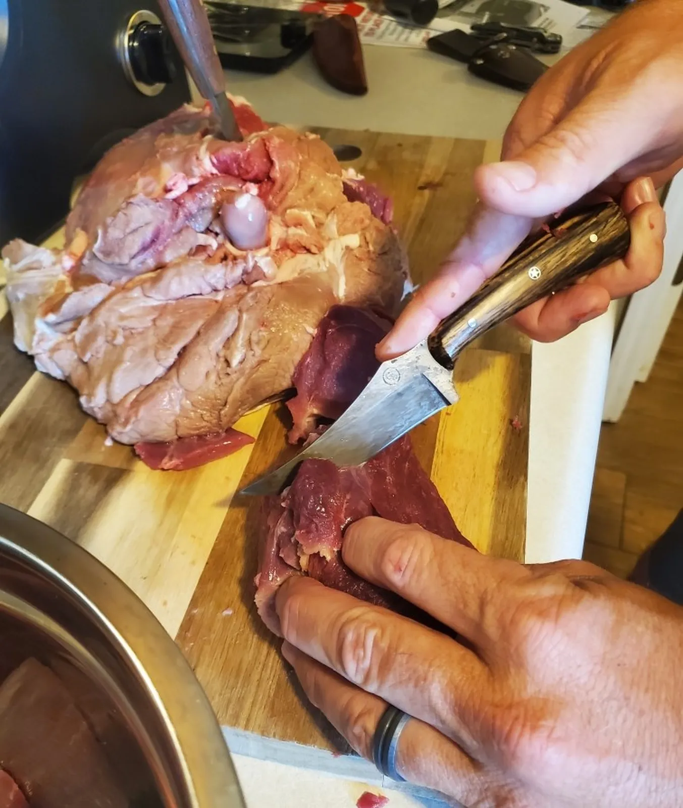 Spalted pecan trailing point knife in use cutting meat, with a red cedar handled three-finger skinner in the background.