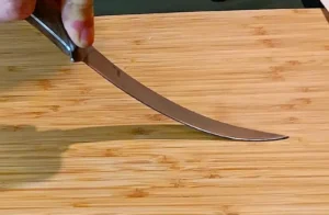 A man's hand gripping the wood handle of a 1095 high carbon steel fillet knife, bending the blade to showcase its flexibility.