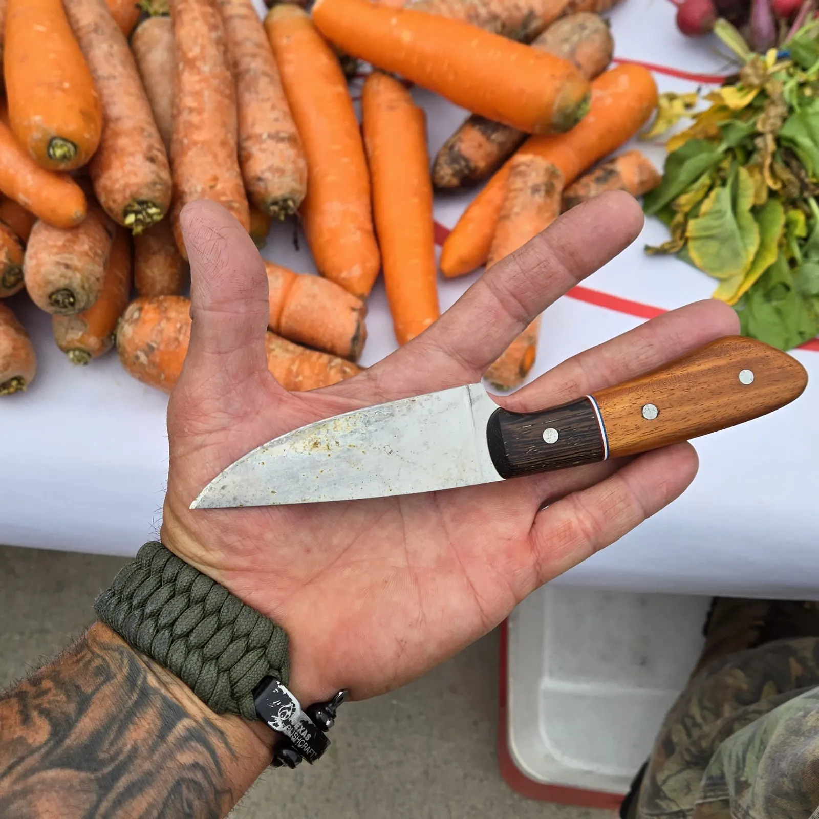Local farmer proudly showing off his new custom knife from GreBec Knives, held in his hand with a pile of fresh carrots behind it.